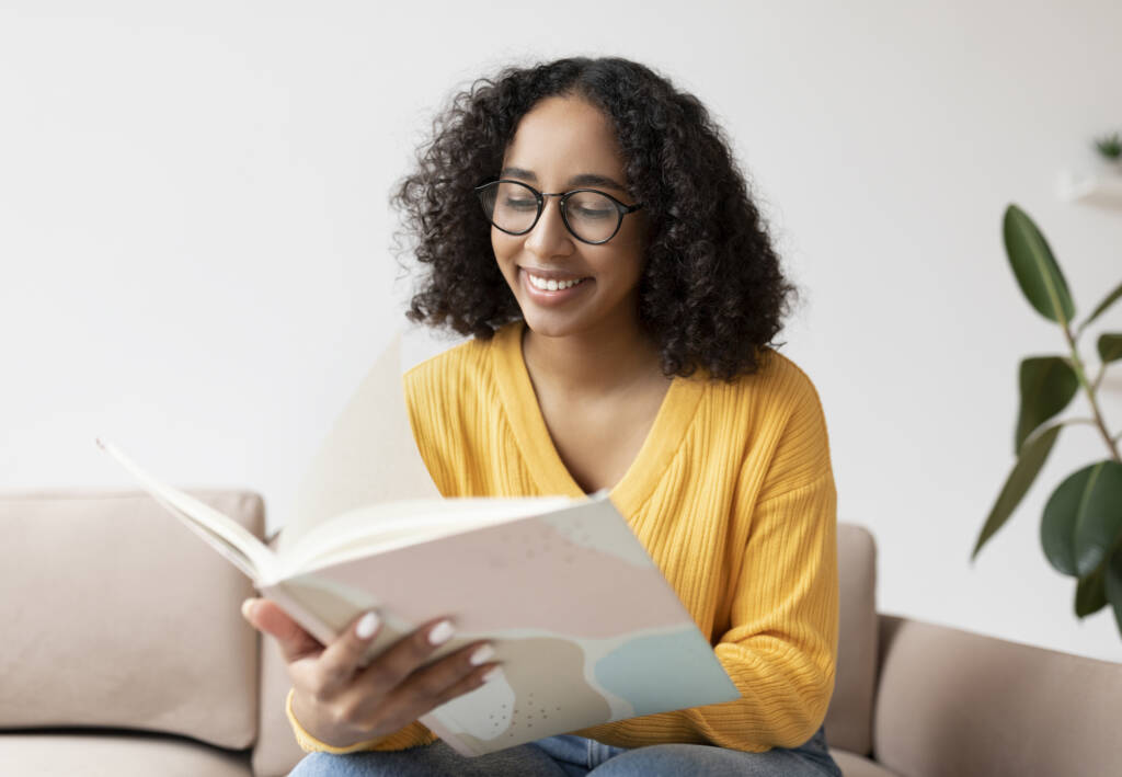 woman in glasses reading book on couch at home.
