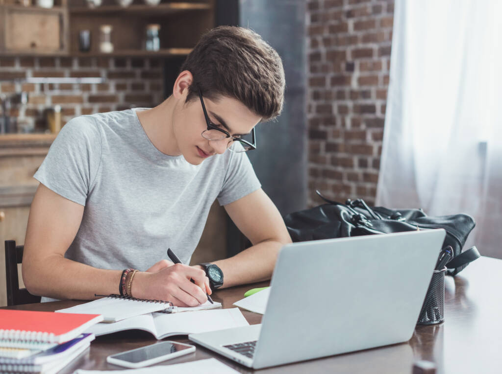 student writing homework at table with laptop and smartphone