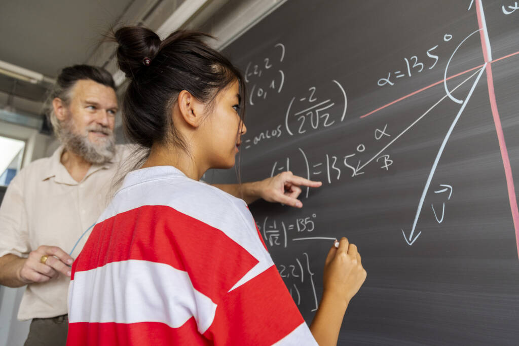 high school maths teacher explains blackboard exercise to student.