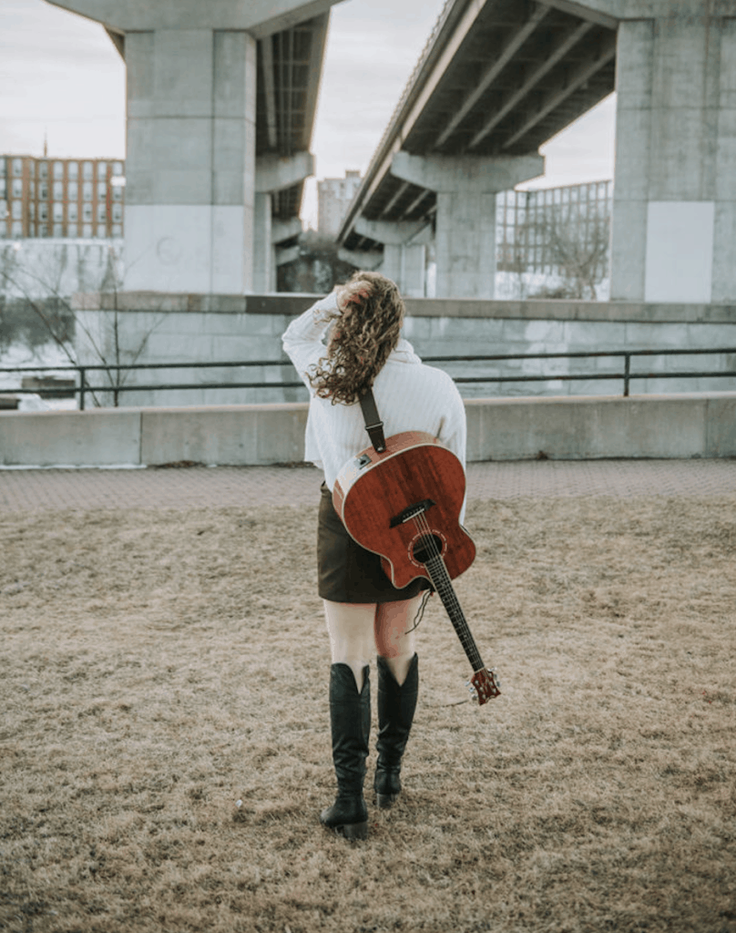 female musician walking under bridge with guitar on her back