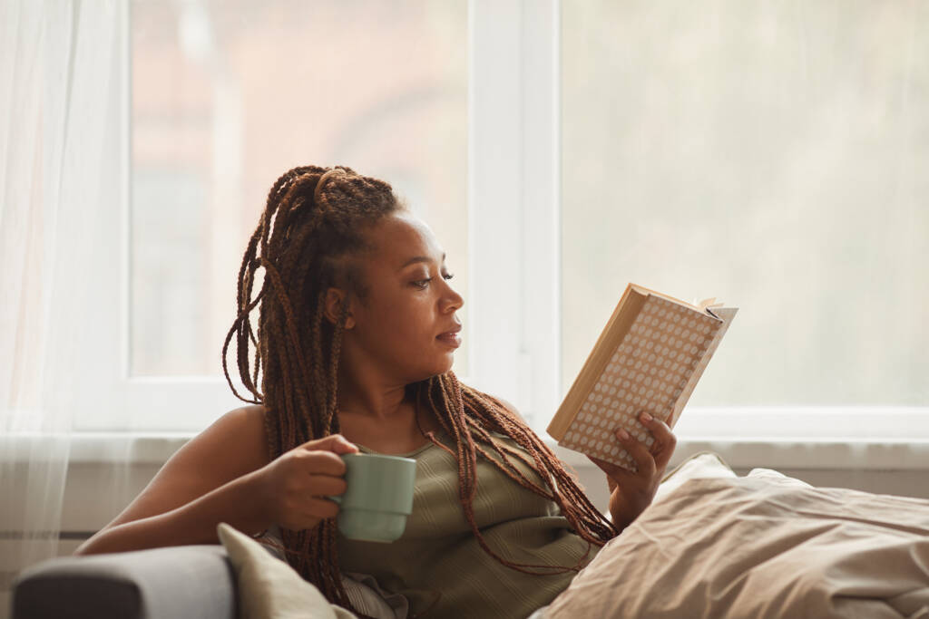 woman lying on sofa drinking coffee and reading an interesting book at home
