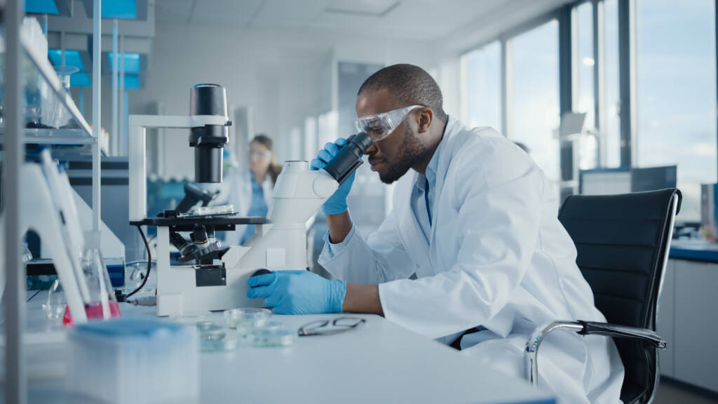 Medical Development Laboratory: Portrait of Black Male Scientist Looking Under Microscope, Analyzing Petri Dish Sample.
