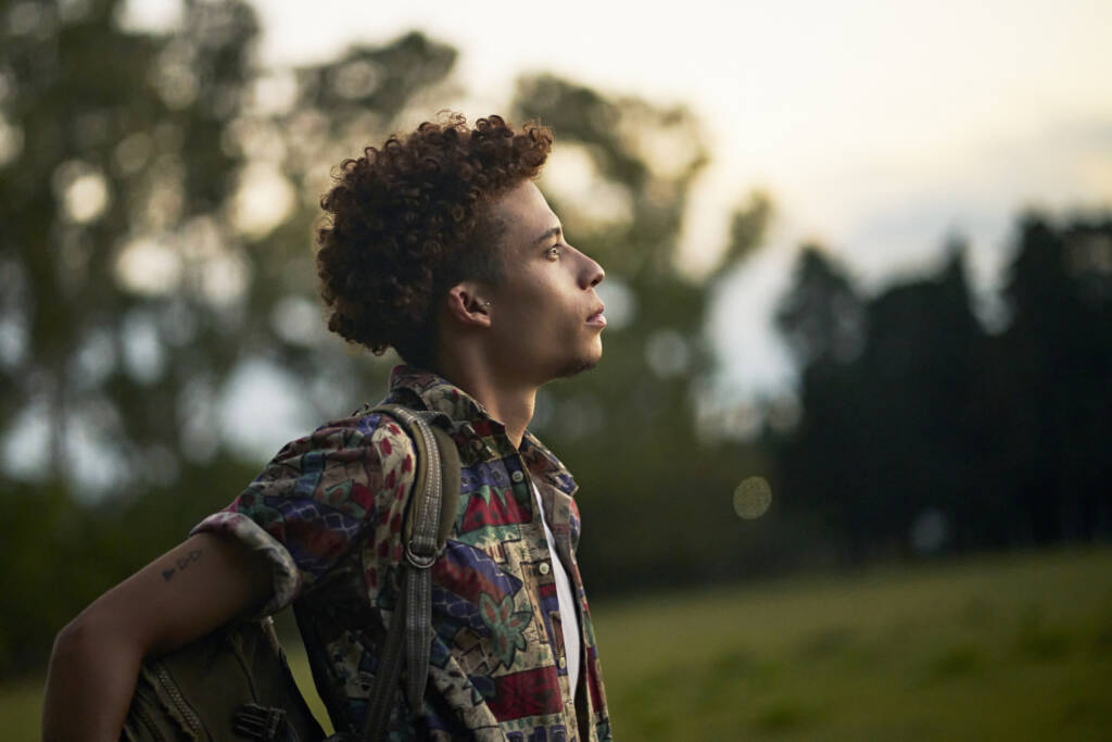 profile view of man with curly brown hair wearing casual clothing and backpack looking away from camera with contented expression of hope.