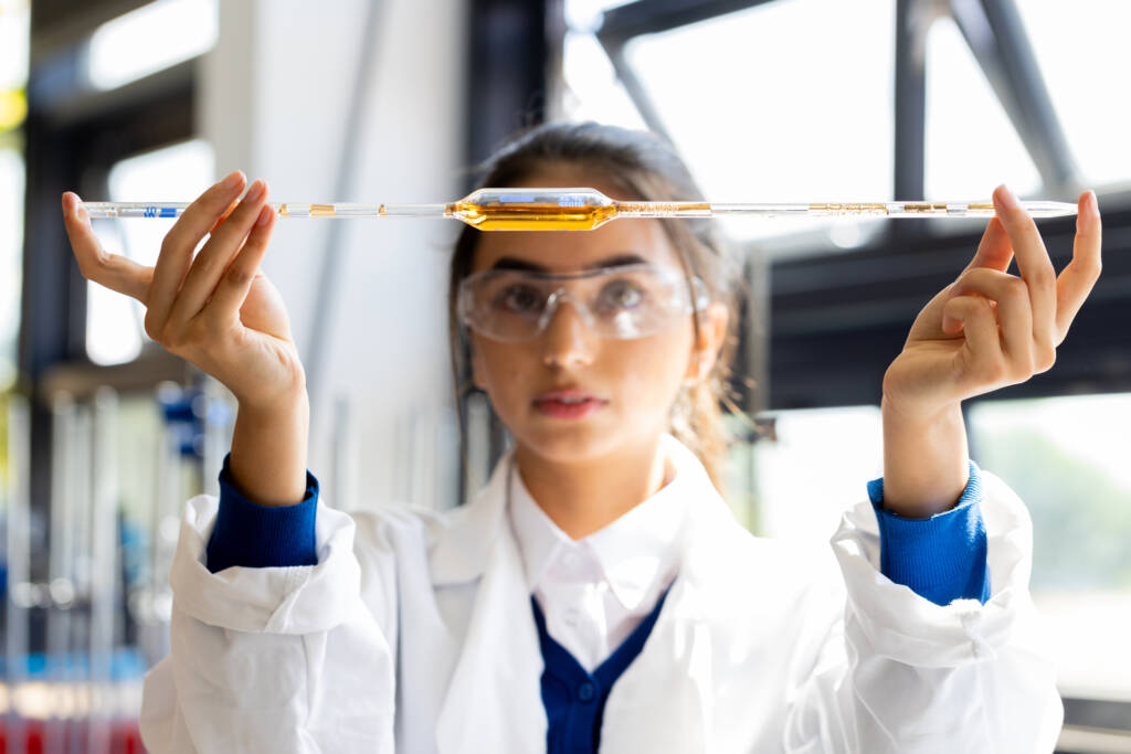 teenager holding up a pipette filled with an orange substance. She is wearing a lab coat and protective eyewear.