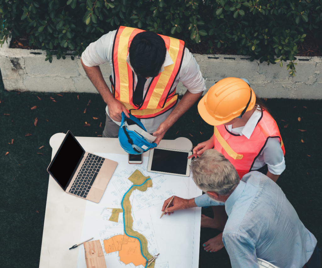 Three construction workers review blueprints and plans at a table outdoors.